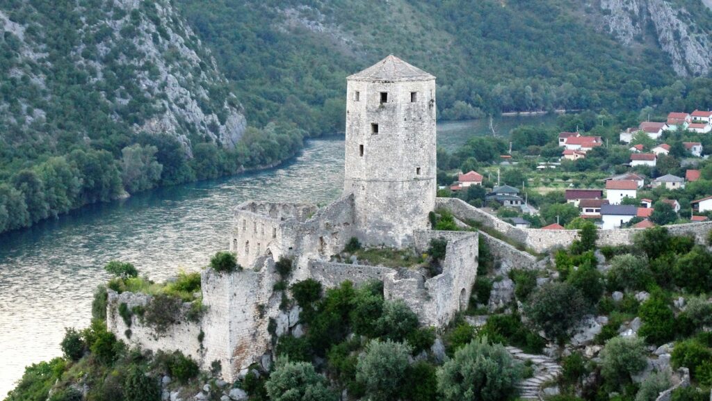 Ancient stone castle by the Neretva River in Bosnia, surrounded by lush greenery and mountains.