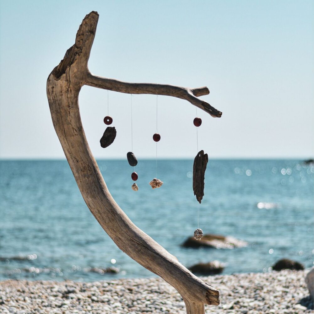 A driftwood sculpture with hanging ornaments on a pebbled beach by the sea.